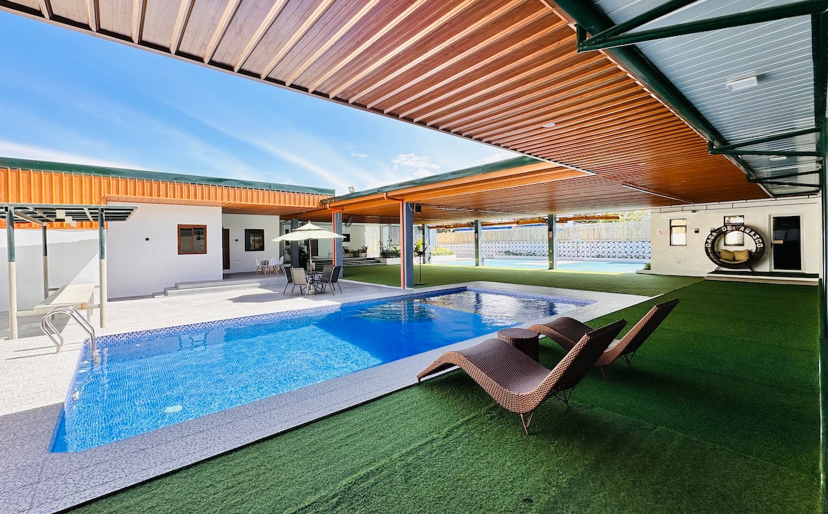 A spacious outdoor area is highlighted by a pool surrounded by a clean, manicured lawn. Lounge chairs are positioned beside the pool, and shaded seating is available under a covered patio, creating an inviting space for gatherings and relaxation.