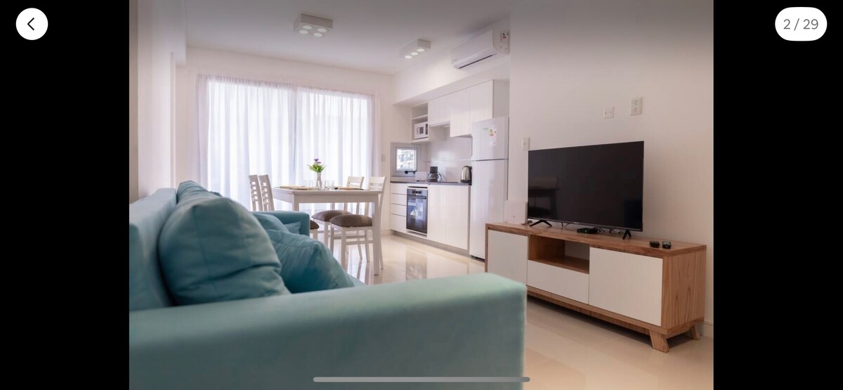 A modern living area is seen featuring a light blue sofa and a dining table set for four. A sleek kitchen with white cabinetry and stainless steel appliances is visible in the background, complemented by large windows allowing ample natural light.