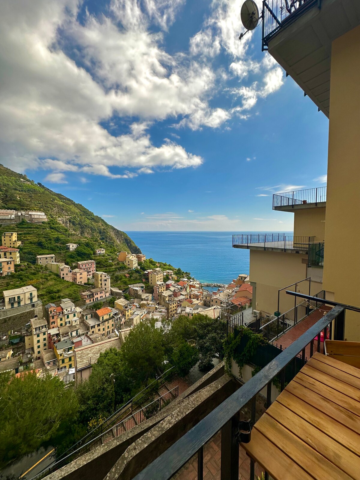 A scenic balcony view captures stunning vistas of the Cinque Terre coastline, showcasing colorful buildings nestled against a lush hillside. The shimmering blue sea stretches into the horizon under a partially cloudy sky, framed by a contemporary balcony railing and wooden furniture.