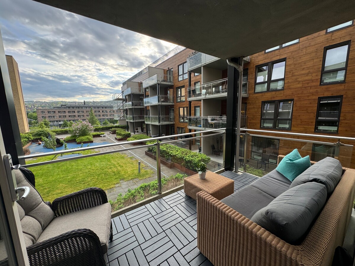 A balcony area is presented with a comfortable sofa and a stylish chair. A small potted plant adorns the surface. The view encompasses a green courtyard with landscaped grass and a swimming pool visible in the distance, framed by modern apartment buildings.