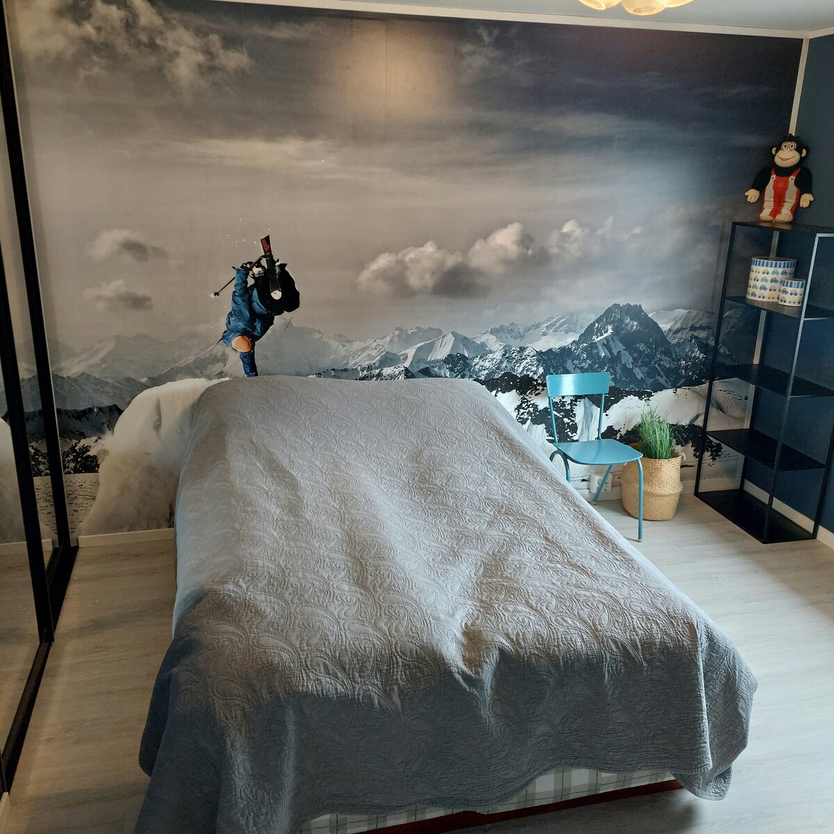 A sleeping area features a spacious bed draped with a grey quilt. Behind, a large mural displays a mountain landscape. A blue chair is positioned nearby, accompanied by a plant in a decorative pot. A black shelving unit stands against the wall.