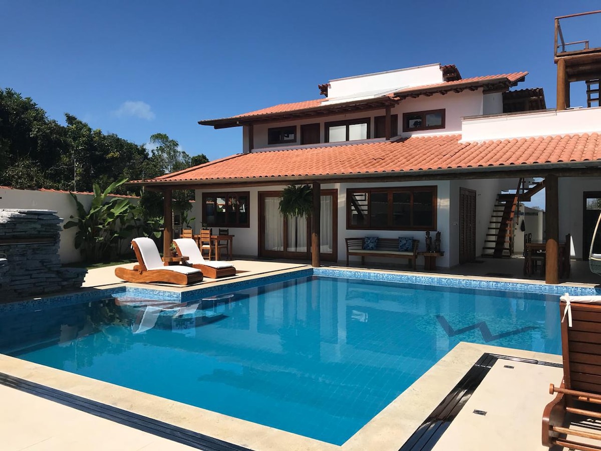 A spacious outdoor area features a large swimming pool surrounded by sun loungers and a wooden dining table. Lush greenery and palm trees create a serene backdrop, while the two-story house showcases a traditional tile roof and large windows, allowing natural light to brighten the interior.
