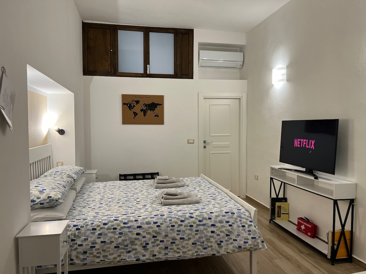 A cozy bedroom features a spacious bed adorned with vibrant, patterned linens. A 43-inch smart TV is mounted on the wall, displaying Netflix. Natural light enters through a small window above, complemented by modern lighting fixtures and an air conditioning unit.