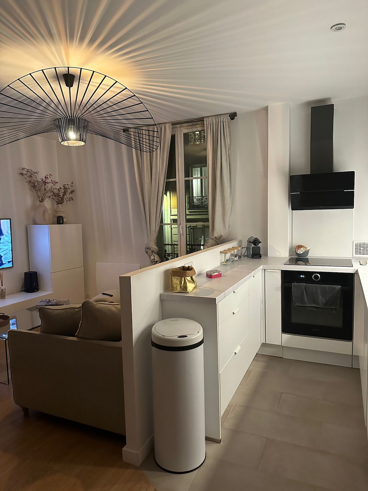 A modern kitchen area is shown, featuring sleek cabinetry and a well-integrated stove. A cozy seating arrangement is nearby, complemented by a decorative light fixture casting soft shadows. Large windows provide natural light, with sheer curtains enhancing the ambiance.