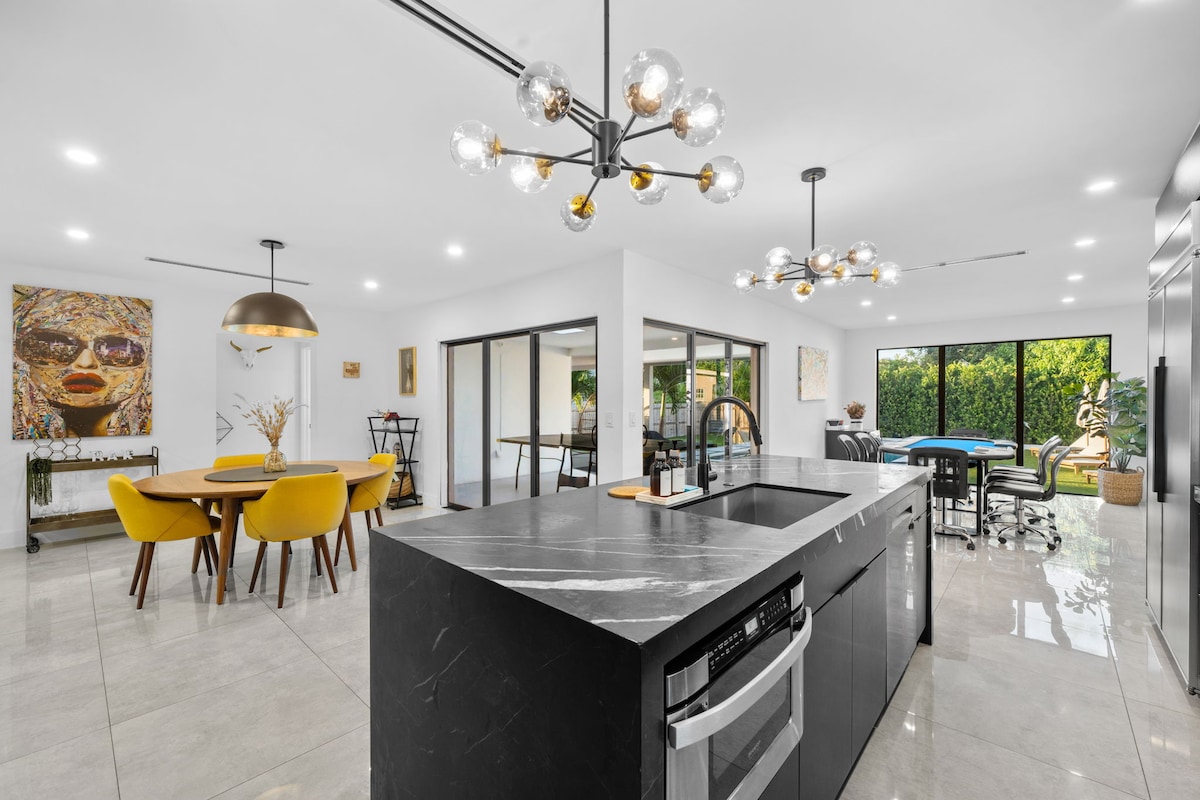 An open-concept kitchen and dining area features a large black stone island with modern appliances. A round dining table with mustard yellow chairs accommodates guests. Large glass doors provide views of the outdoor space, enhancing natural light and creating a seamless transition between indoors and outdoors.