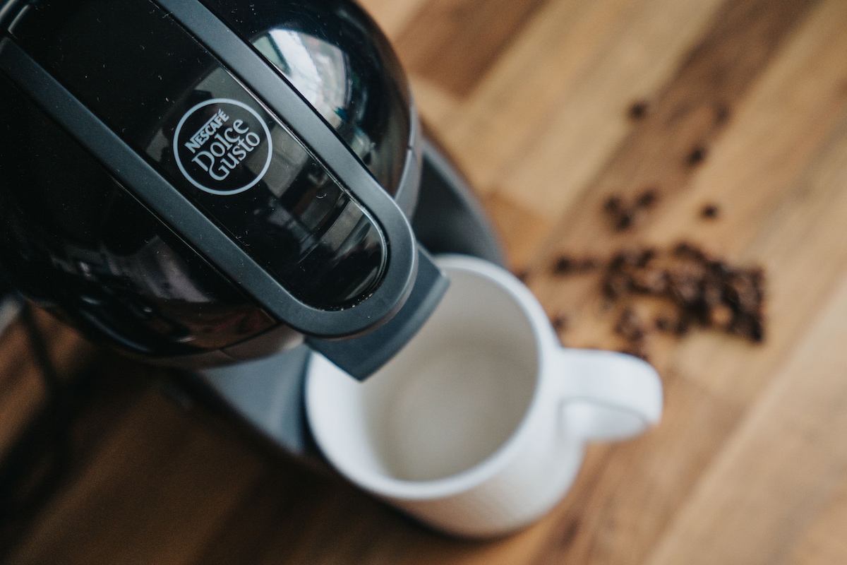 A coffee machine is positioned above a white mug, with coffee beans scattered on the wooden counter. The sleek design of the machine contrasts with the warm tones of the wood, inviting guests to enjoy a fresh brew.