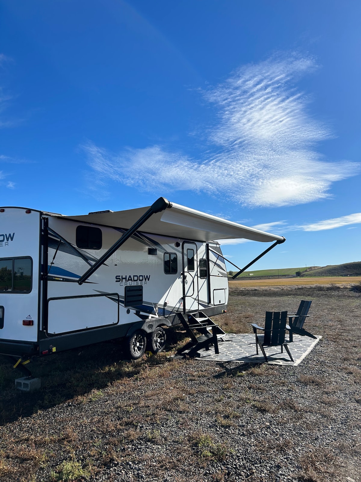 A travel trailer is positioned outdoors, featuring an extendable awning over a patio area with two green chairs. A gravel space surrounds the trailer, with a clear blue sky visible above and rolling hills in the distance.