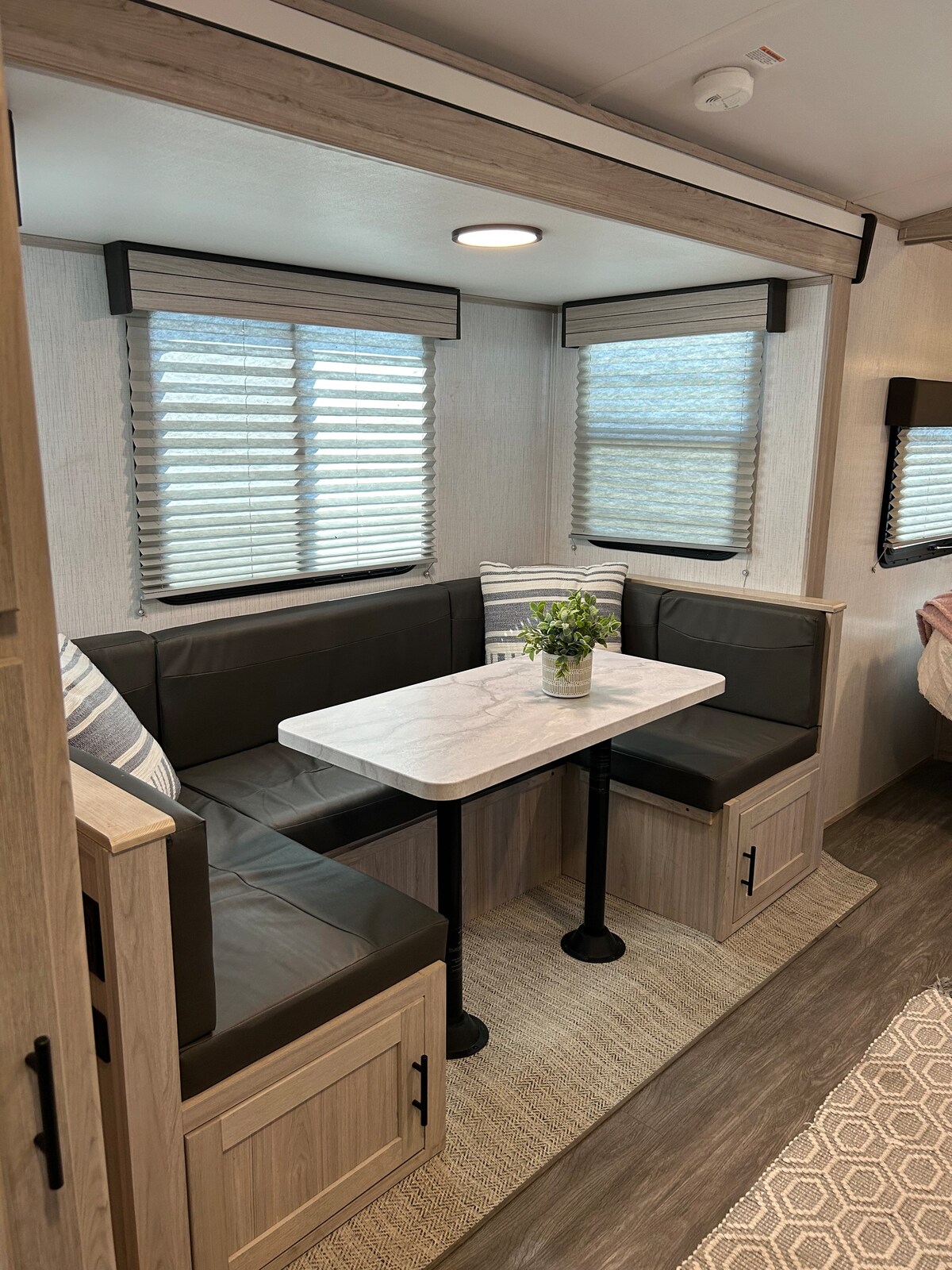 A compact dining area is presented with a marble-topped table surrounded by dark seating. Natural light enters through two windows adorned with horizontal blinds. Light-colored walls and a decorative rug contribute to a clean and organized appearance in the space.