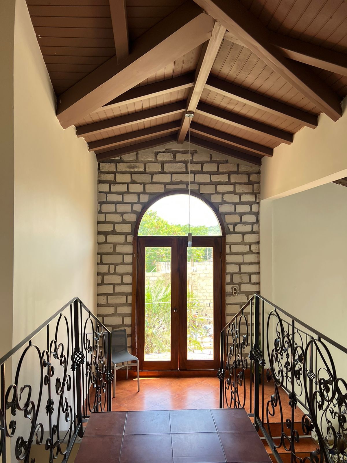 An inviting entrance hall features a high, vaulted ceiling with wooden beams. Decorative railings frame the staircase, leading to large arched doors that open to a view of the outdoors, where greenery can be seen through the glass.
