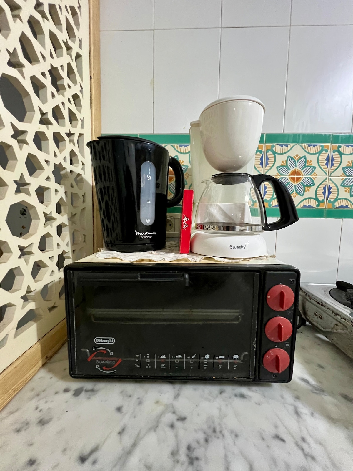 A countertop is occupied by a black electric kettle and a white coffee maker, both positioned beside a compact toaster oven. The appliance arrangement is complemented by decorative tiles featuring a colorful pattern, adding a vibrant touch to the kitchen space.