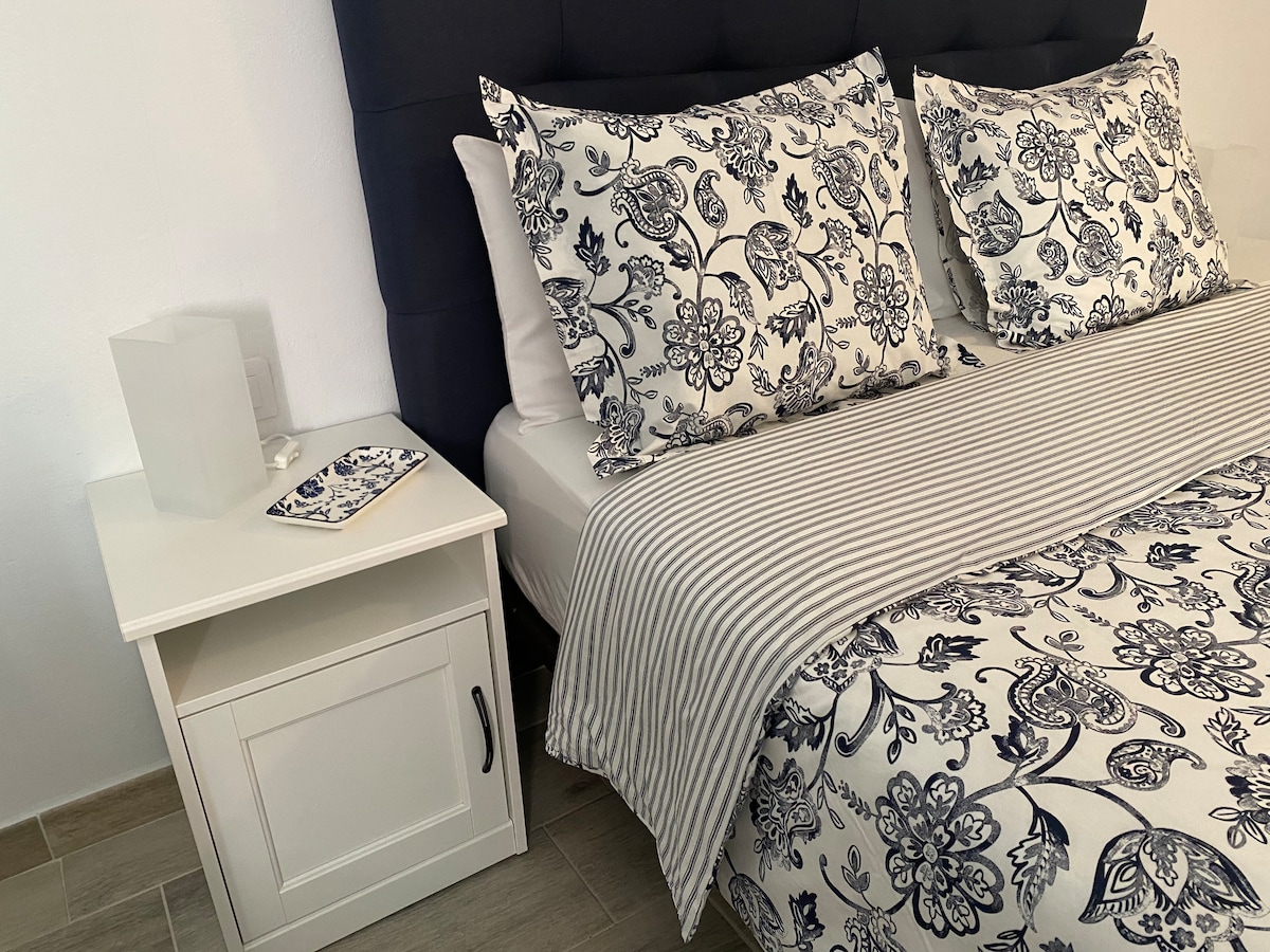 A cozy sleeping area features a spacious bed with patterned bedding and two decorative pillows. A nightstand, with a small decorative plate, is placed beside the bed. Soft lighting from a bedside lamp enhances the relaxing atmosphere.
