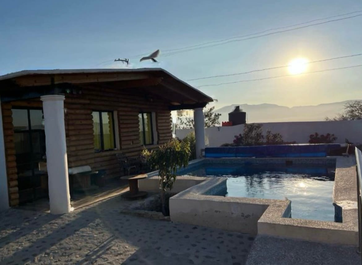 A private villa features a rustic wooden exterior under a clear sky. A serene pool area is visible, reflecting the sun. Surrounding landscaped areas include a small seating arrangement, enhancing the outdoor space for relaxation.