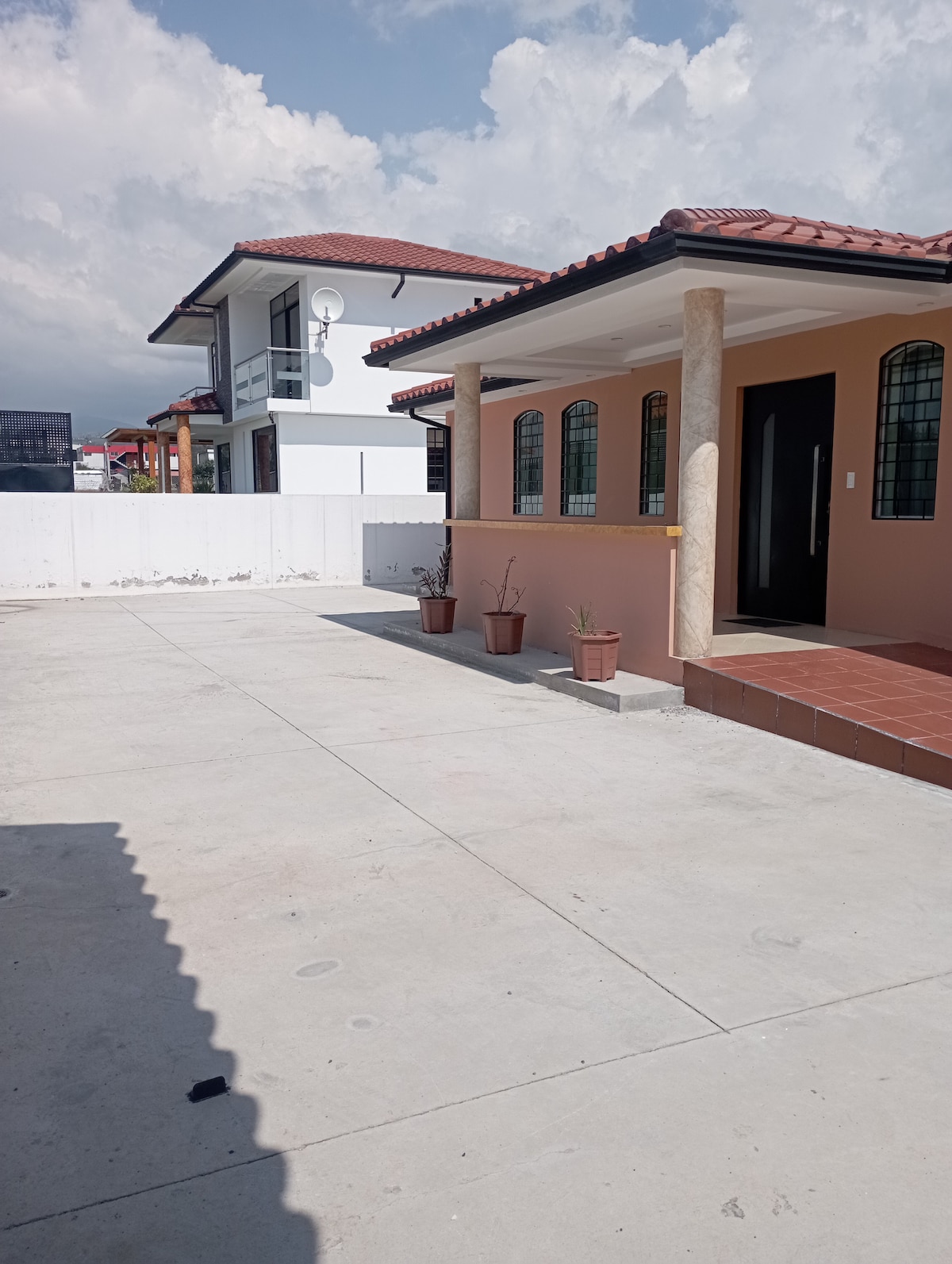 A spacious outdoor area is visible, featuring a smooth concrete surface and two single-story houses in the background. Flower pots with small plants are placed near the entrance of the house on the right. The sky is partly cloudy, creating a bright atmosphere.