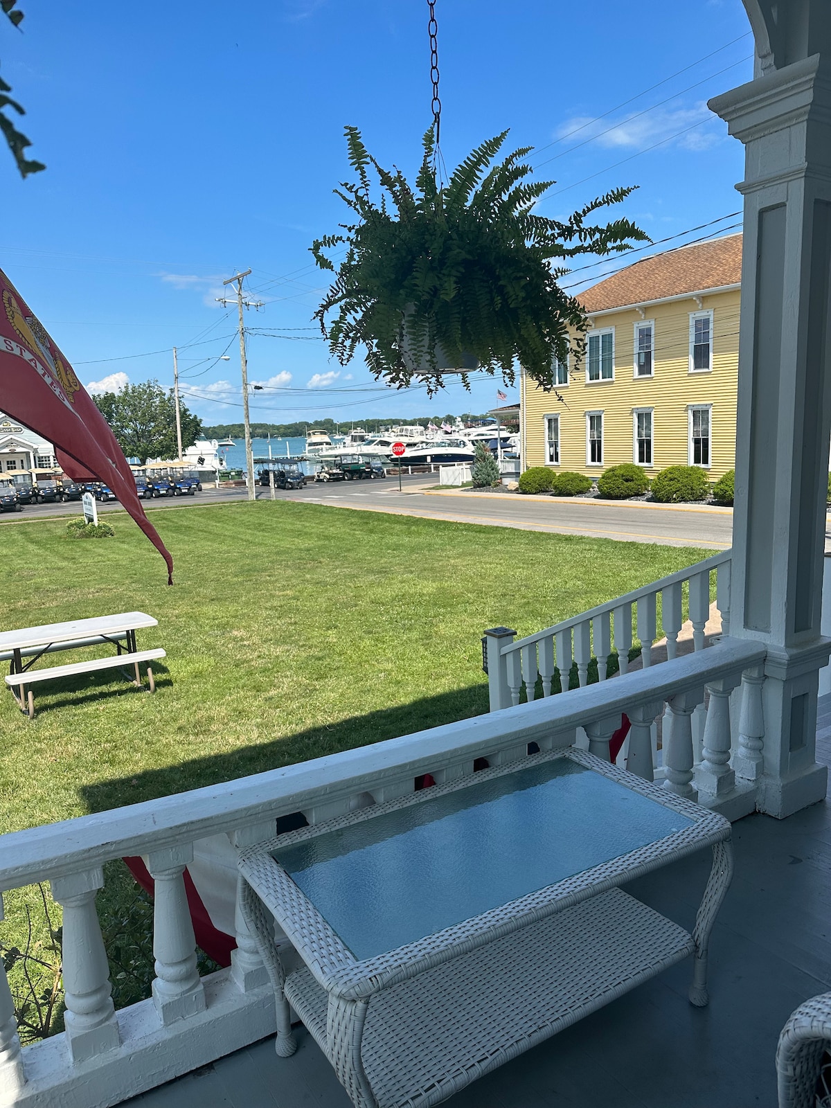 A peaceful outdoor area is visible with a hanging fern planter and a small white table. A grassy lawn extends towards a picturesque bay, while a flag gently waves in the breeze. Nearby buildings reflect a charming coastal community.