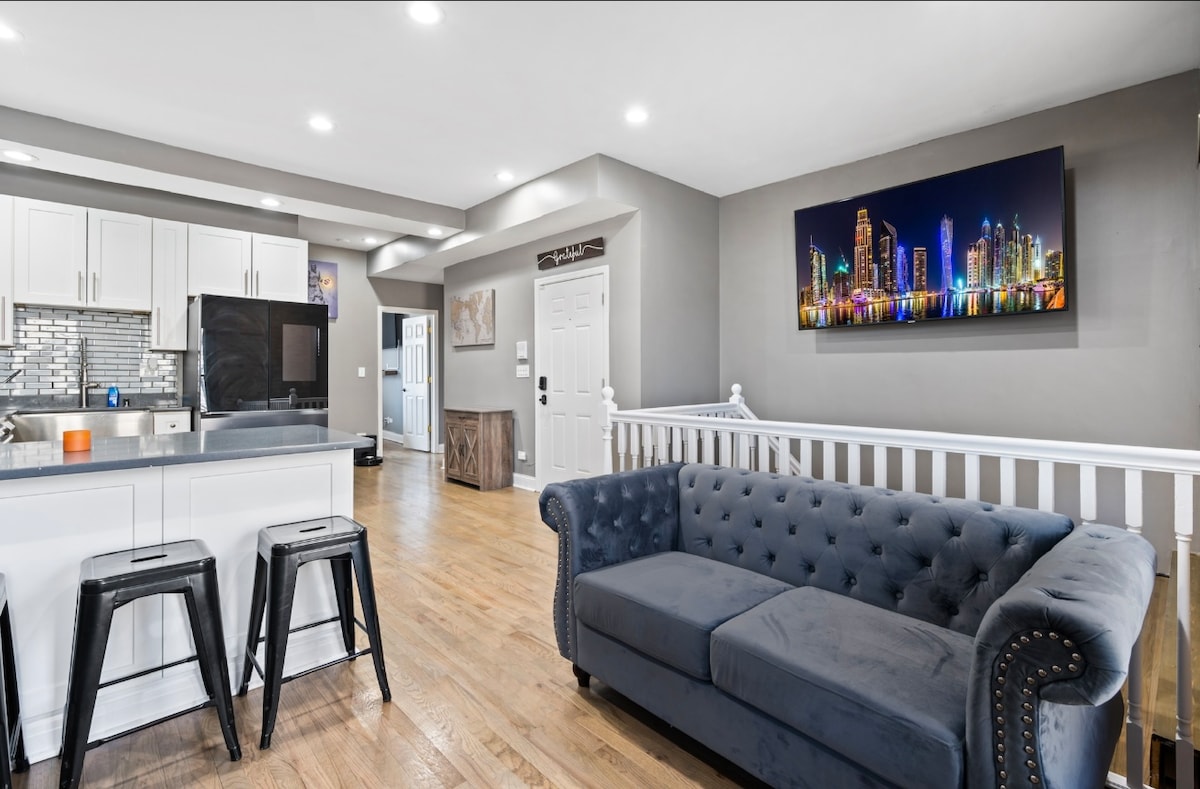 The open kitchen and living area feature modern cabinetry, stainless steel appliances, and a navy blue tufted sofa. A colorful panoramic cityscape photo hangs on the wall above, while sleek black stools provide seating along the counter. Natural light enhances the warm wooden flooring.