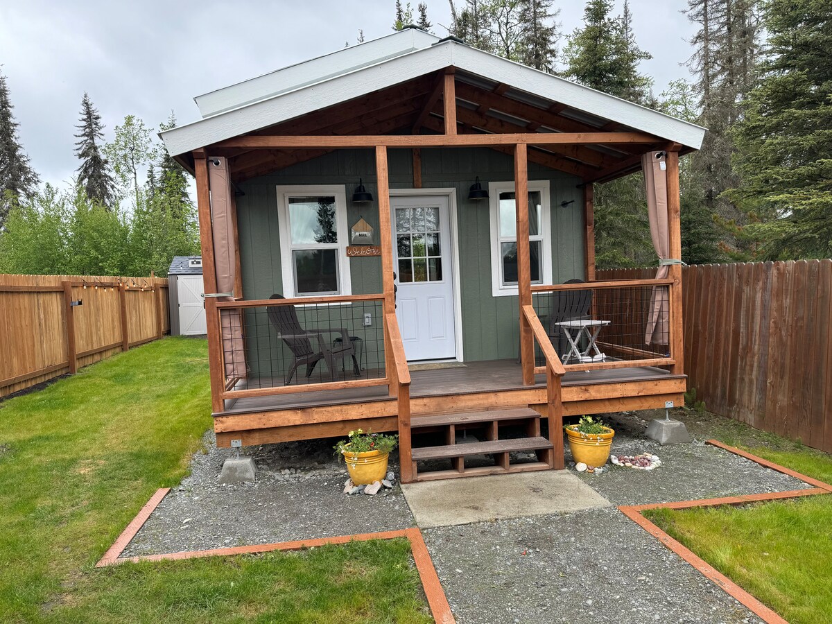 The exterior of a charming tiny home features a covered porch with two chairs and a small table. Potted plants flank the entrance. A spacious yard with well-maintained grass and a gravel path leads to steps, while a wooden fence provides privacy.
