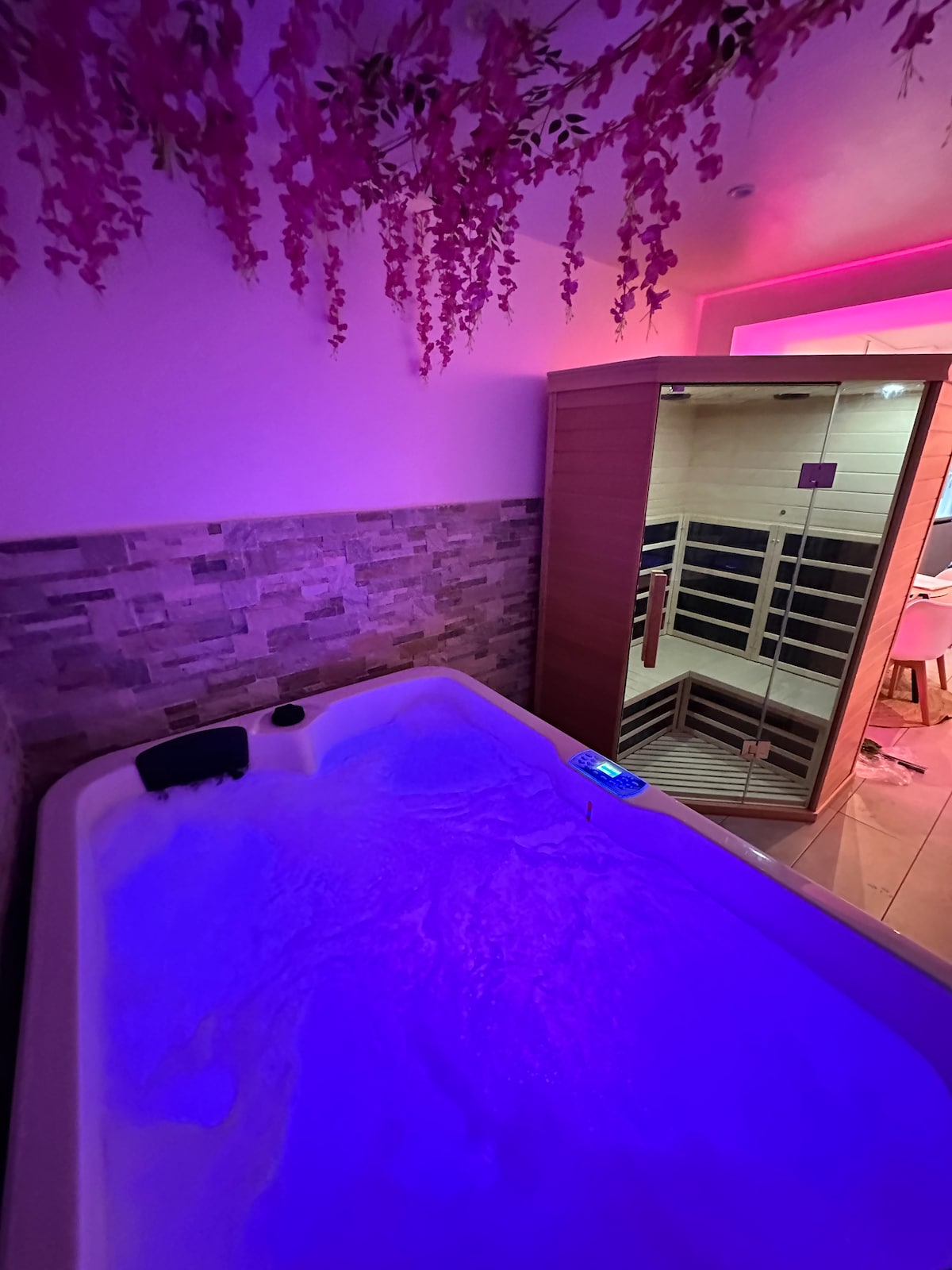 A private jacuzzi and sauna are presented, illuminated by soft, colored lights. The jacuzzi features bubbling water and is surrounded by textured stone walls and decorative foliage above. The sauna, made of wood, provides a serene and cozy ambiance for relaxation.