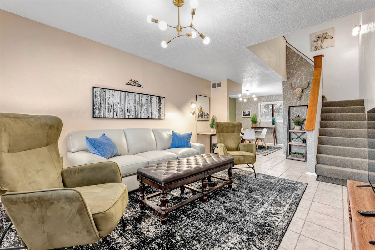 A spacious living area is presented with a light-colored sofa and two armchairs on a decorative rug. A coffee table is centered, and the walls are adorned with art. Stairs in the background lead to the upper level, while a dining area is visible in the distance.
