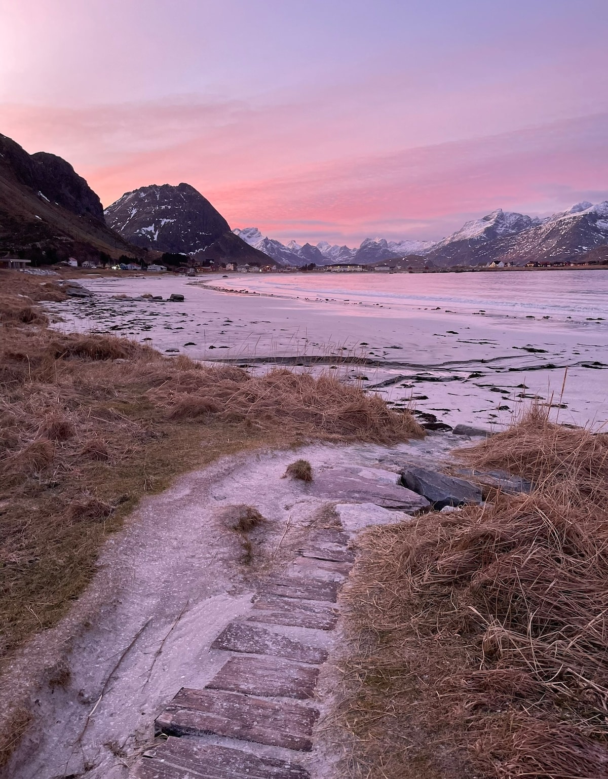 A scenic landscape is captured, featuring a winding path leading through tall grasses along a serene beach. The shoreline is bordered by snow-capped mountains, and a soft pastel sky reflects the colors of sunset over the calm waters.