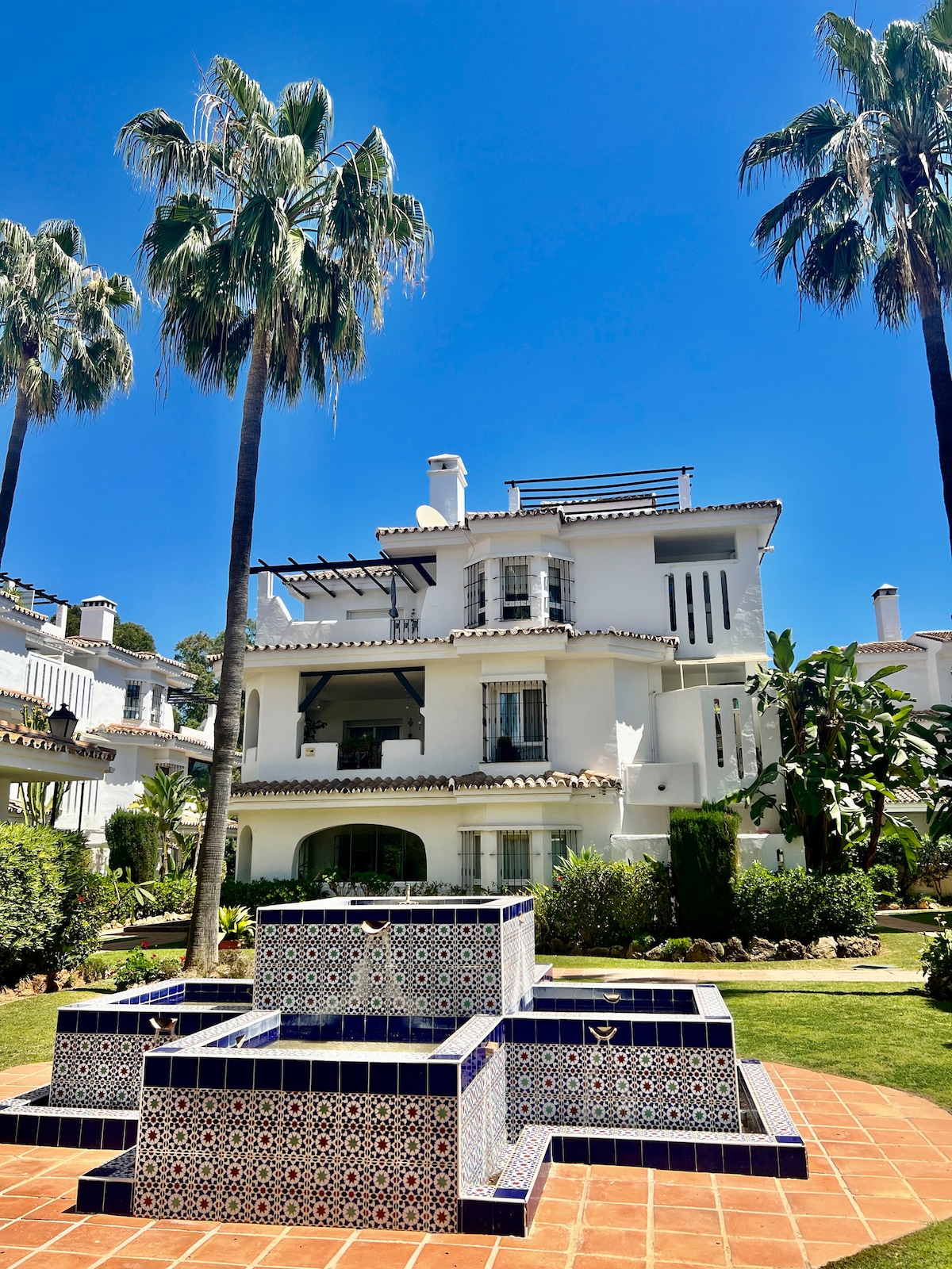 A three-story Andalusian-style penthouse is surrounded by lush gardens. The courtyard features a decorative fountain with intricate blue and white tile work. Tall palm trees create a tropical ambiance under a clear blue sky, emphasizing the tranquil and serene setting.
