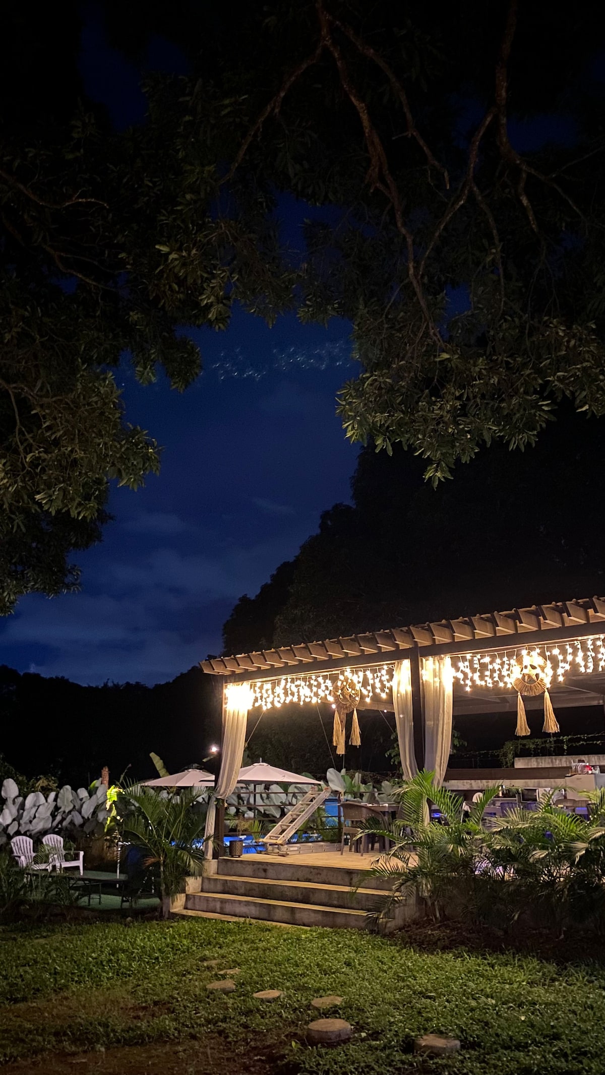 A gazebo is illuminated by soft string lights, creating a warm ambiance in the evening. Surrounding greenery provides a natural backdrop, while shadows of trees add depth to the scene. The setup suggests a welcoming space for communal gatherings and outdoor dining.
