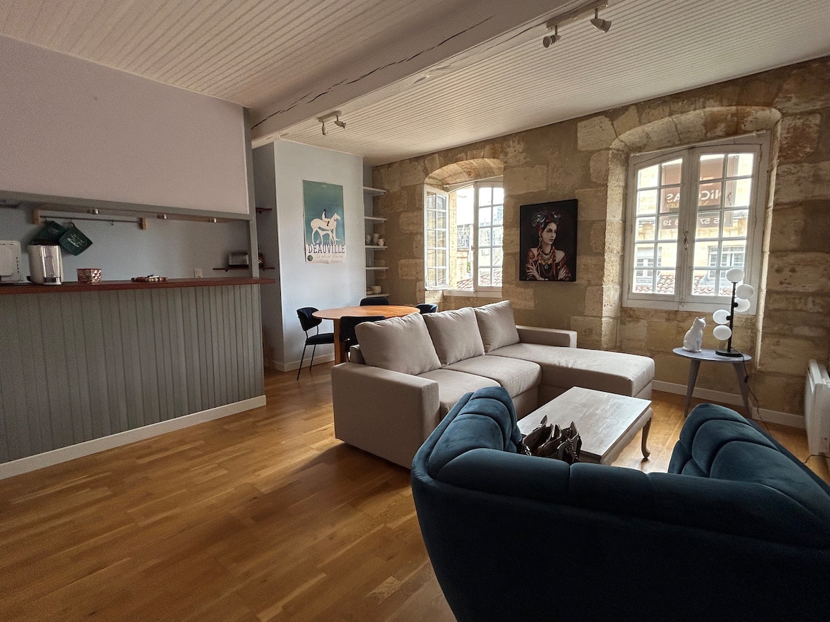 A spacious living area is featured, showcasing a corner sofa and an armchair. Large windows allow natural light to illuminate the wooden floor. A dining table is visible in the background, complemented by a kitchenette area, blending functionality and comfort.