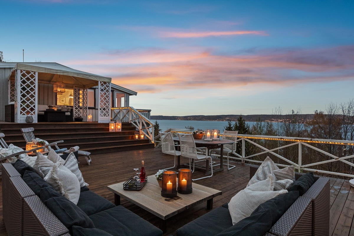 A spacious terrace is seen with comfortable seating, decorated with soft cushions. Several tables are set with candles, creating a warm ambiance. The background highlights a scenic view of the water, complemented by a sunset sky with hues of pink and orange.