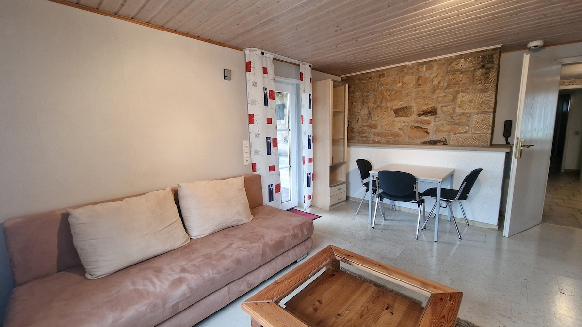 A cozy living area features a brown sofa with plush cushions, complemented by a wooden coffee table. A small dining table with chairs occupies one side, with a light-colored stone wall adding texture to the space. Natural light enters through a door with patterned curtains.