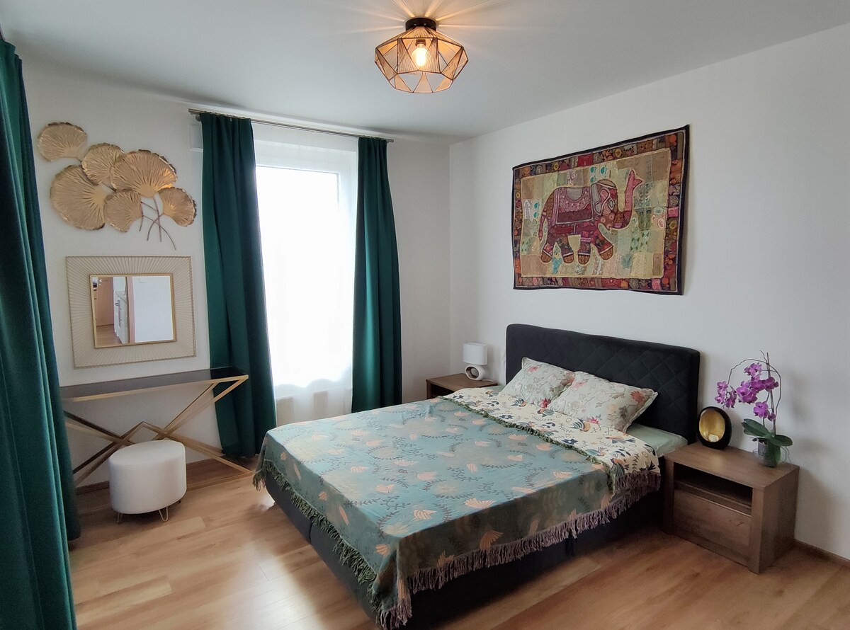 A well-appointed bedroom features a spacious bed adorned with a decorative blanket. Surrounding elements include a mirror and a small accent table. A colorful wall tapestry and potted orchid enhance the decor. Natural light filters through a window, complemented by green curtains.