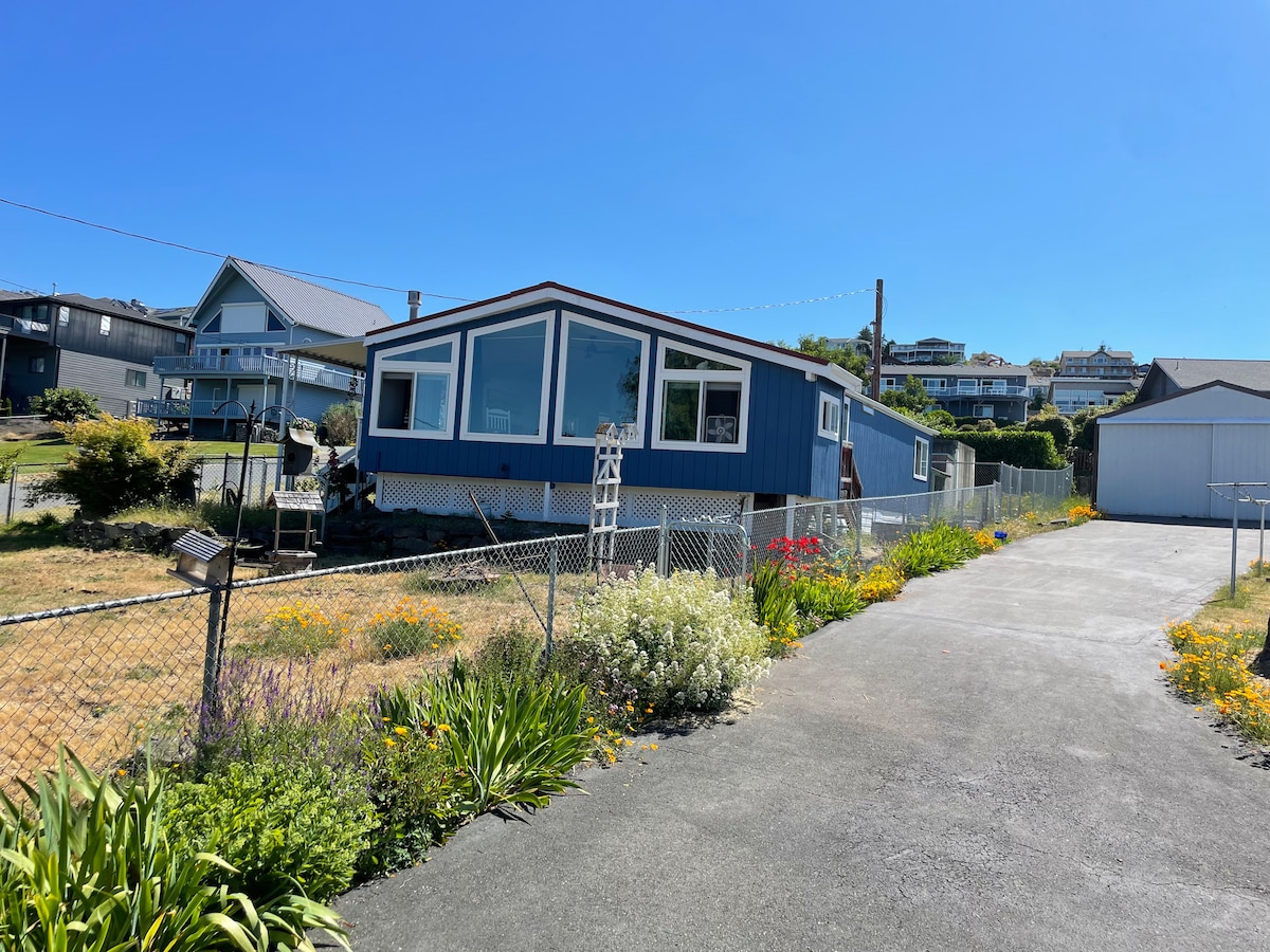The exterior of the home is shown, featuring a blue exterior with large windows. A well-maintained garden with colorful flowers borders the property. The paved driveway leads to a nearby structure, and the fenced yard provides a sense of privacy.