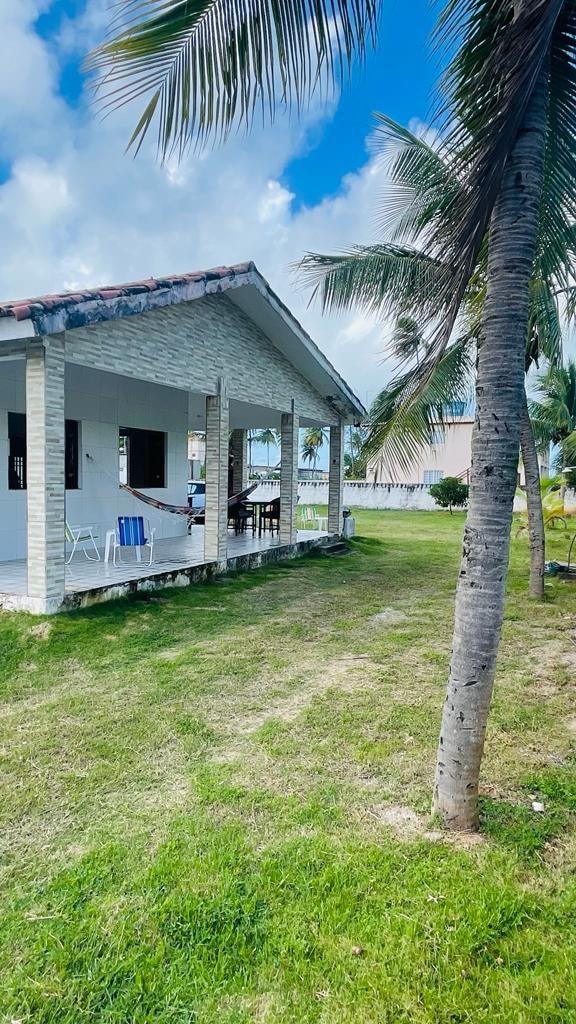 A spacious exterior view features a light-colored house with a sloped roof, surrounded by lush green grass and palm trees. The covered porch provides seating options, creating a relaxed outdoor area for guests. Bright blue skies and scattered clouds add a serene backdrop.