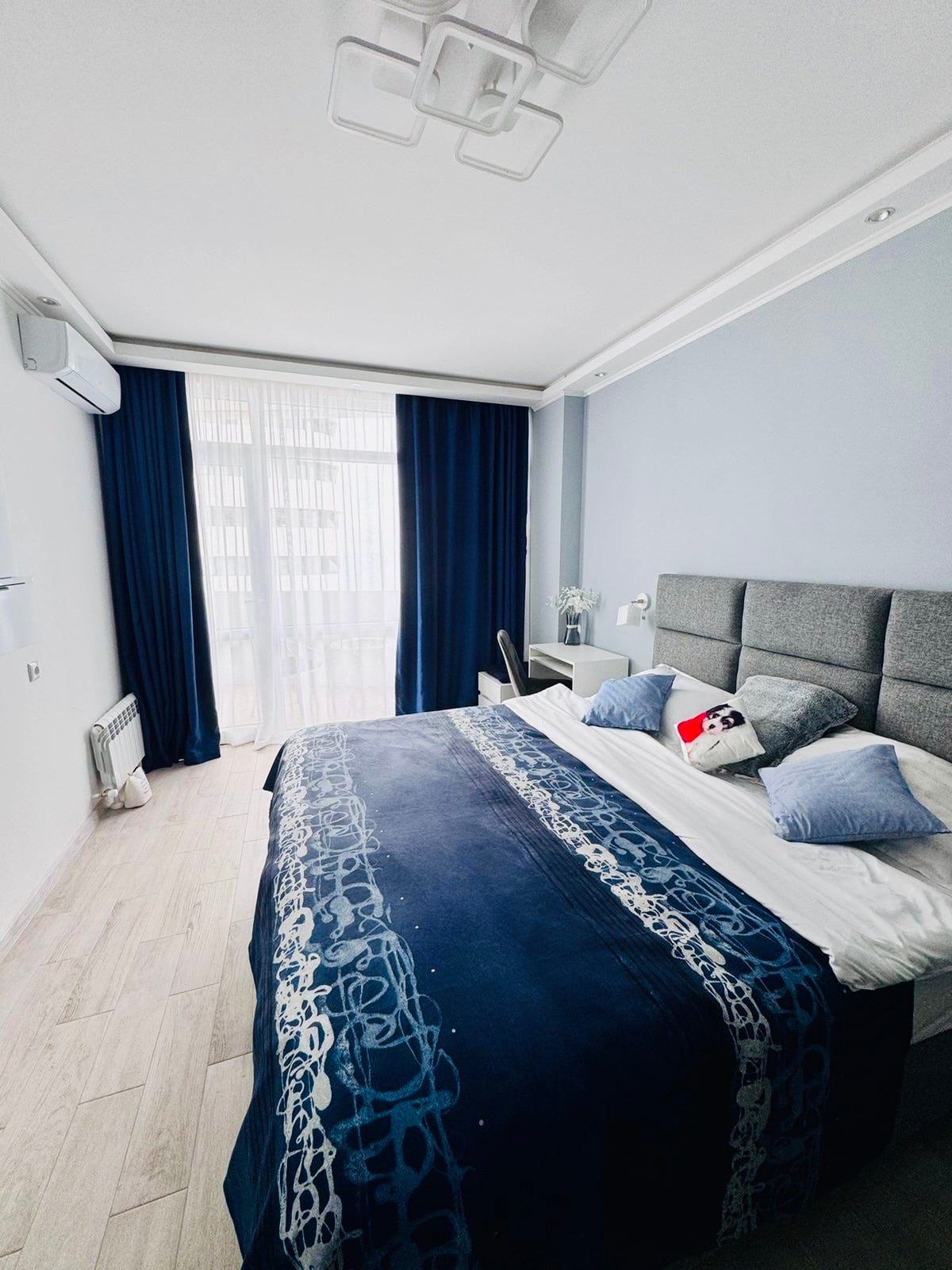 A modern bedroom is equipped with a spacious bed featuring a blue patterned coverlet. Light filters through sheer curtains, enhancing the room's brightness. A small desk and chair complement the space, while air conditioning is mounted on the wall for comfort.