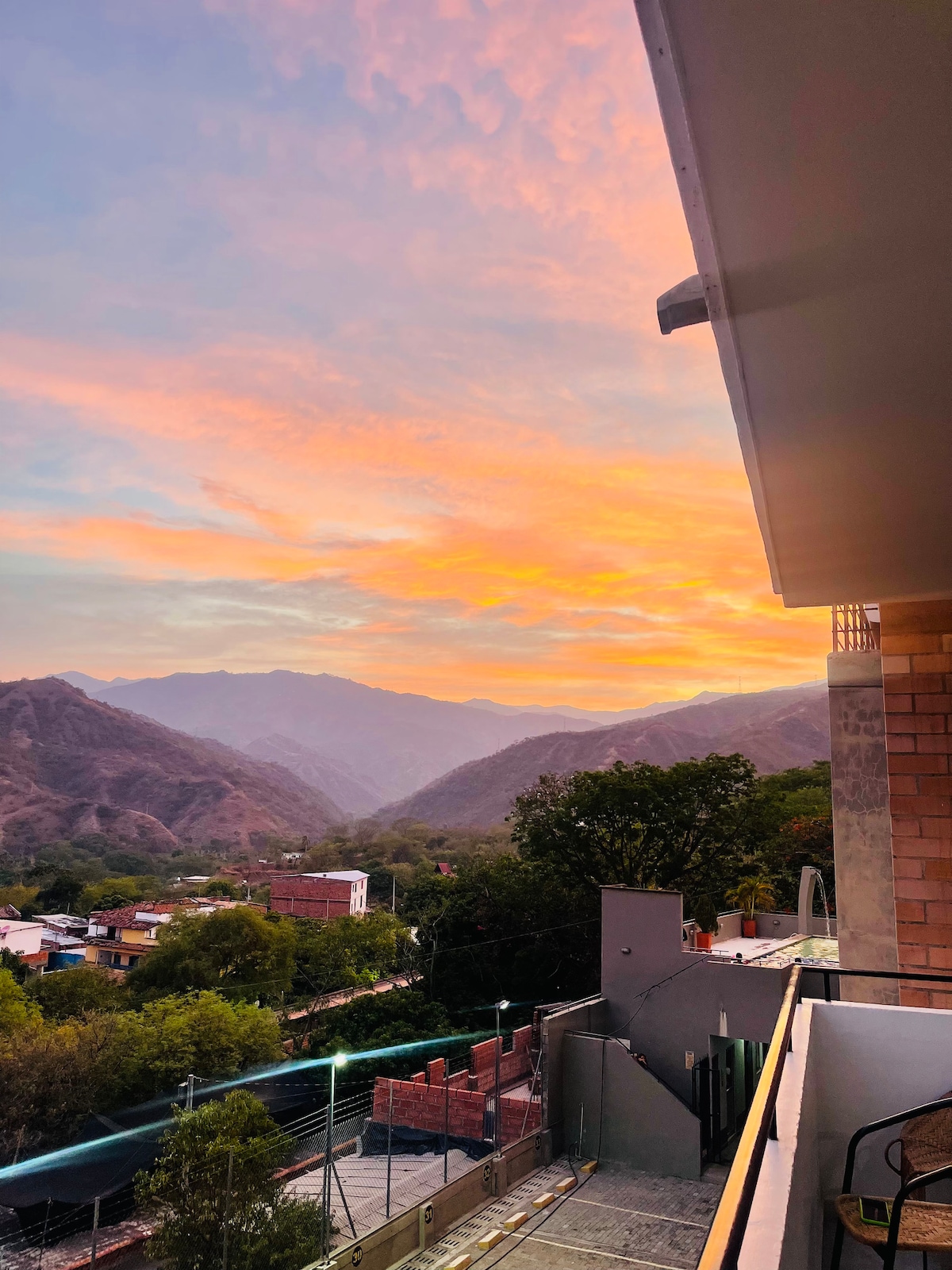 A picturesque view stretches across the landscape, featuring rolling mountains bathed in warm hues of orange and pink during sunset. The balcony railing is visible, framing the natural beauty beyond, while trees and rooftops can be seen in the foreground.