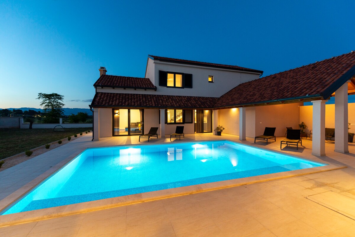 A spacious outdoor area features a well-lit pool with a serene turquoise color, surrounded by lounge chairs. The two-story house with a tiled roof stands in the background, its windows softly glowing in the evening light, creating a welcoming ambiance.