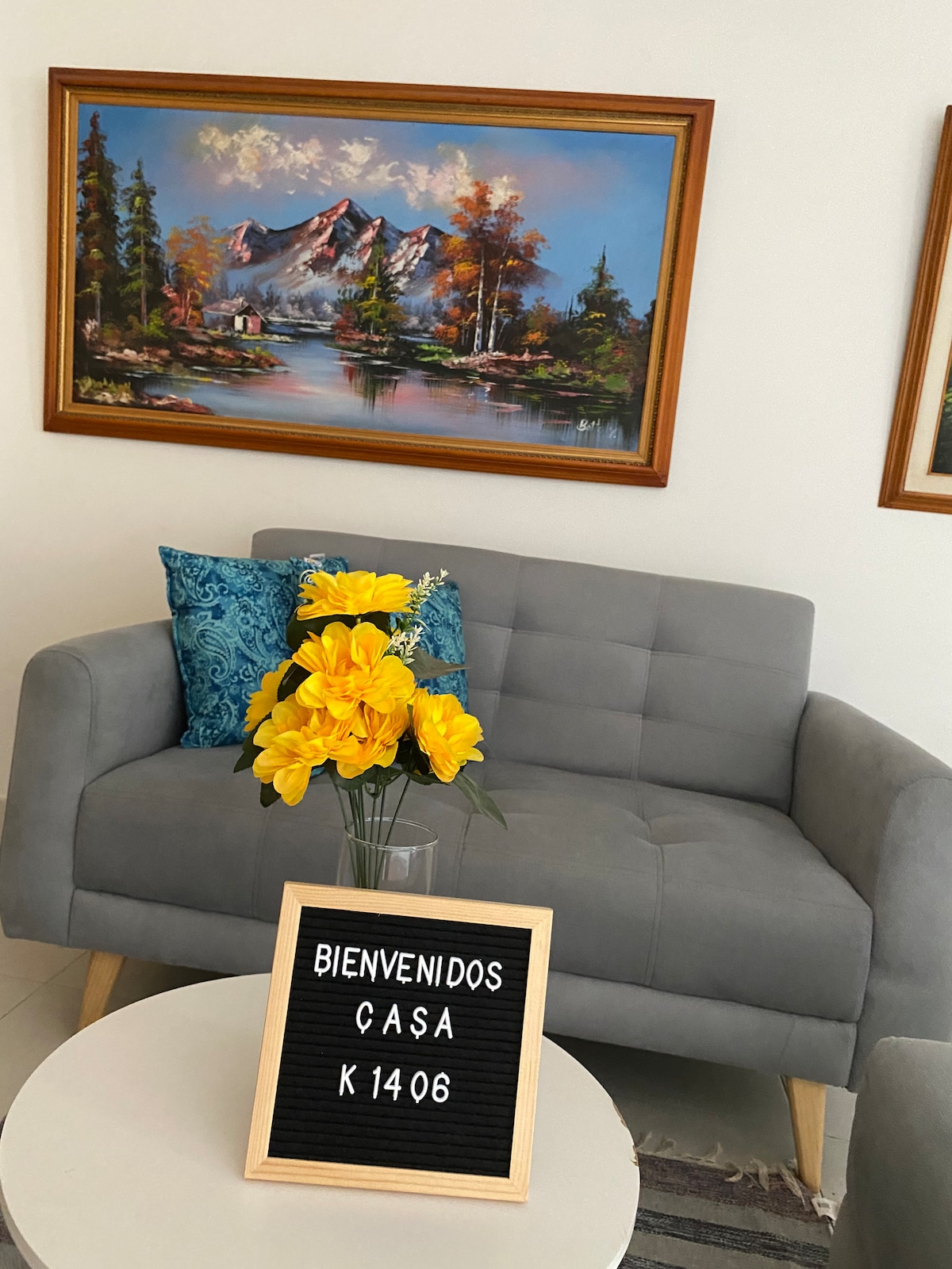A comfortable gray sofa is positioned in front of a vibrant mountain landscape painting. A small round table displays a welcome sign, and a vase with yellow flowers adds a touch of color to the arrangement.