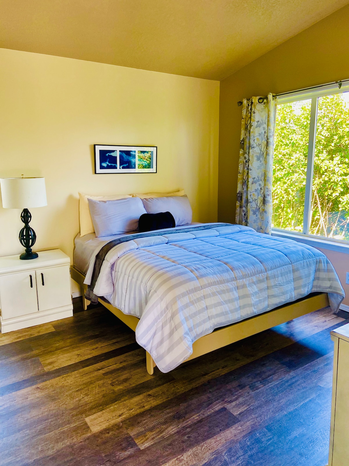A bright bedroom is presented with a spacious bed covered in soft bedding, positioned near a large window that brings in natural light. A bedside lamp and a light-colored nightstand complement the decor. Bright green foliage is visible outside the window.