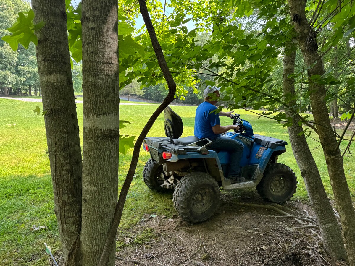 An all-terrain vehicle is positioned on a grassy area, surrounded by trees. A person is seated on the ATV, facing away from the camera. The scene conveys a connection with nature, with vibrant green grass and shaded tree trunks visible.