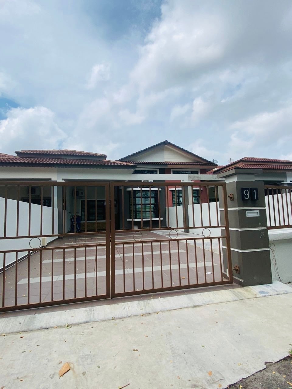 A welcoming entrance is featured with an ornate gate and a pathway leading to the front door. The house's exterior combines warm tones with a tiled roof, and large windows allow natural light to illuminate the area. The property is enclosed by a boundary wall.