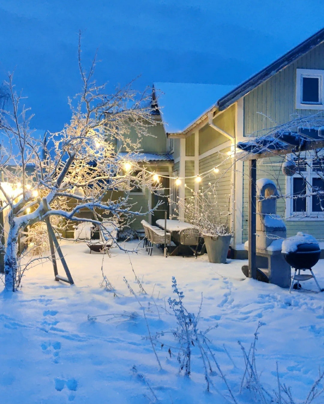 A serene courtyard is blanketed in snow, with soft evening light illuminating the scene. Outdoor furniture is arranged for dining, and string lights create a warm ambiance among the frosted trees. A grill and a swing add charm to the peaceful winter landscape.