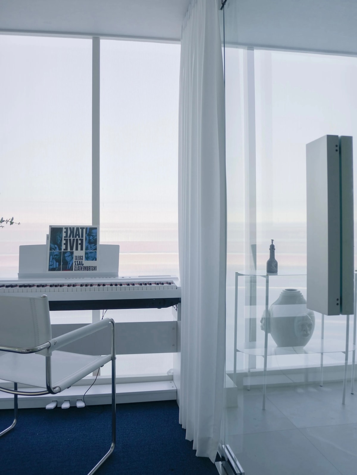 A modern workspace is highlighted, featuring a white piano with sheet music placed atop. A clear glass partition separates the area, creating an open ambiance. The sleek design includes a white chair and minimalist decor, with sheer curtains allowing soft light to filter through.