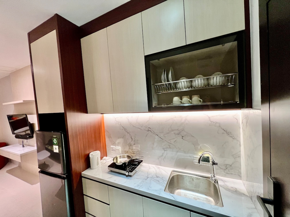 A modern kitchen area features sleek cabinetry with a marble countertop and an under-cabinet light. A stainless steel sink is positioned next to a gas stovetop, with white dishware arranged in a glass-front cabinet above. A refrigerator is located on the left.