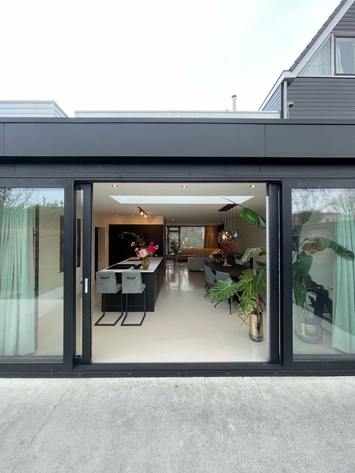 An inviting view of an open-concept living space is presented through large glass doors. The area features a modern kitchen with bar seating and a spacious living room, complemented by decorative plants and floral arrangements.