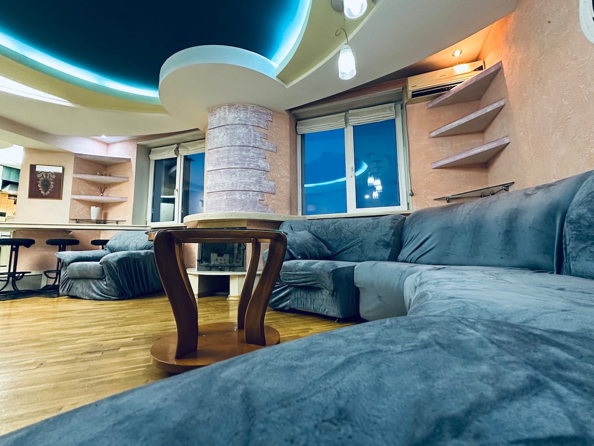 A spacious living area features a large, comfortable sofa adorned with soft textiles. A circular wooden table stands in the center, surrounded by light-colored shelving. Multiple windows allow natural light to illuminate the room, with softly glowing ceiling illumination adding a modern touch.