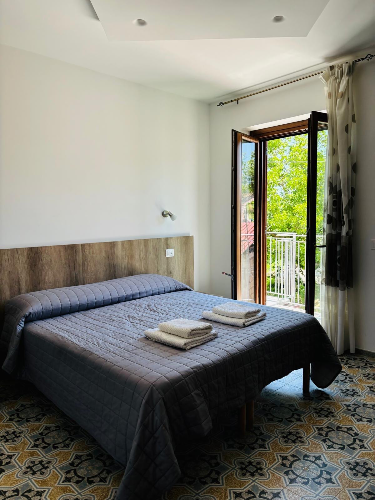 A cozy bedroom features a spacious bed covered with a textured gray bedspread. Two rolled towels are neatly placed at the foot of the bed. Natural light filters through a partially open window that leads to a small balcony, offering views of greenery.
