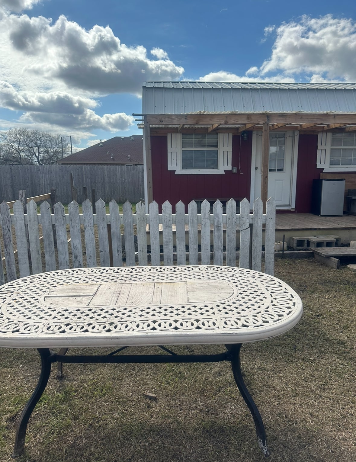 A white metal table with intricate cutout designs is positioned in front of a small red cottage. The cottage features a tin roof and white-framed windows. A picket fence surrounds the outdoor area, and a clear blue sky with fluffy clouds is visible above.