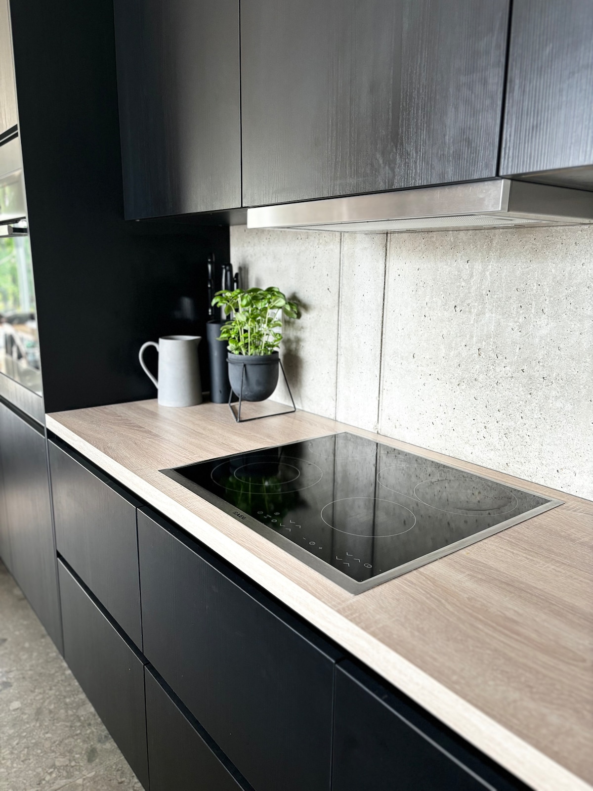 A modern kitchen scene features sleek black cabinetry and a light wooden countertop. An induction cooktop is visibly integrated into the counter, while a small potted plant adds a touch of greenery. Gentle light reflects off the surfaces, creating a clean and contemporary look.