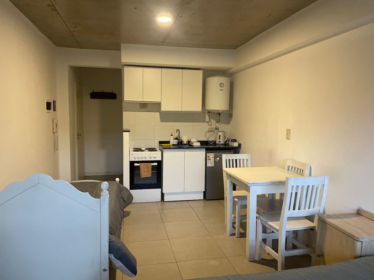 A compact kitchen area features white cabinetry and a gas stove. A dining table with four chairs occupies the space, while a gray sofa is visible to the side. The tiled floor complements the overall neutral color scheme, with natural light illuminating the room.