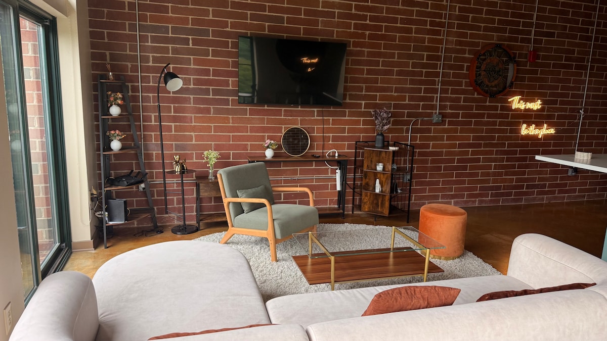 A stylish living area features a light sofa facing a wall-mounted television. A modern armchair and a small, circular orange stool complement the space. A glass coffee table sits atop a soft area rug, while brick walls provide a warm backdrop, accentuated by decorative plants and wall art.