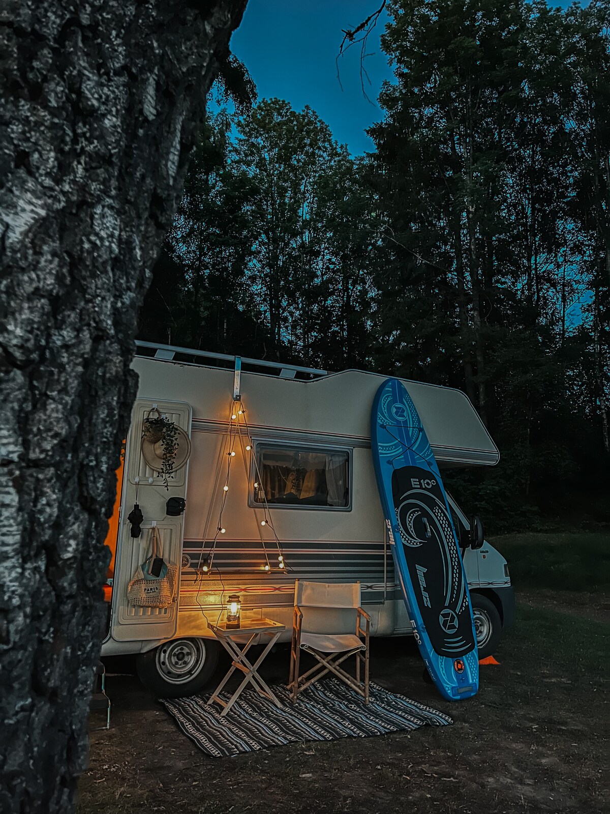 The motorhome showcases an inviting outdoor setup with fairy lights overhead, a small table, and two folding chairs positioned on a textured rug. A paddle board leans against the side of the vehicle, surrounded by a natural forest backdrop.