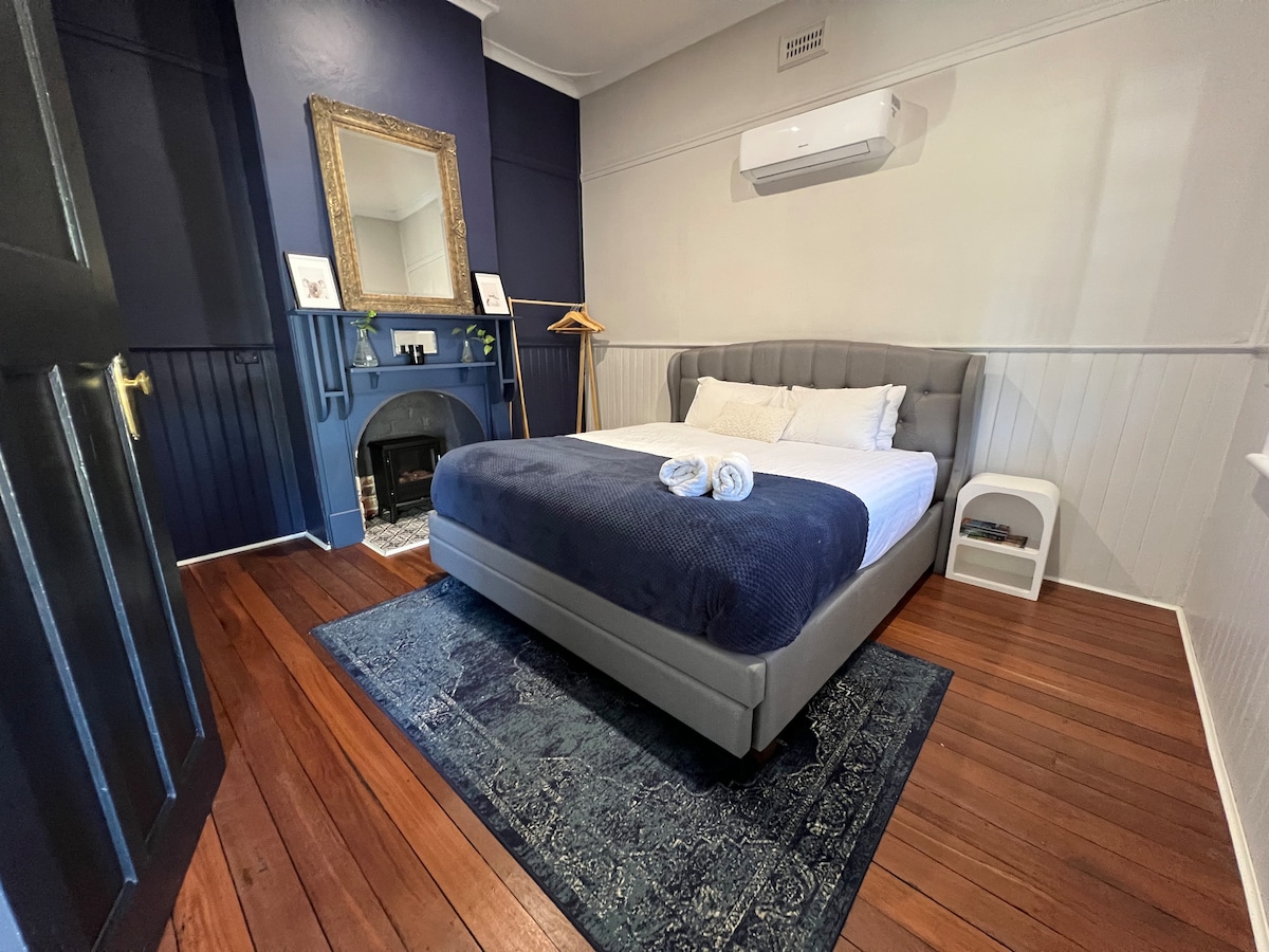 The master bedroom features a spacious bed with a dark blue throw blanket, flanked by two towels. A vintage-inspired fireplace is visible beneath a large mirror, and natural light enters from a window. Original jarrah floorboards complement the room's neutral and navy walls.